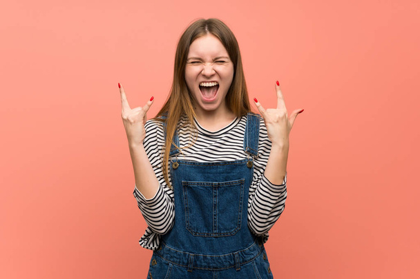 Young woman with overalls over pink wall making rock gesture - Photo, Image