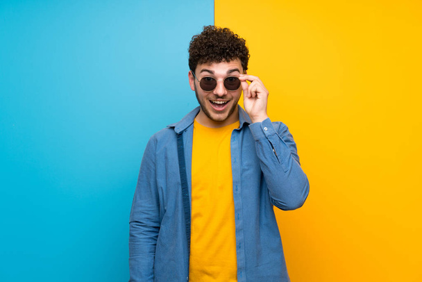 Hombre con el pelo rizado sobre la pared de colores con gafas y sorprendido
 - Foto, Imagen
