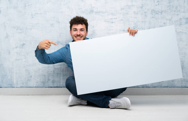 Genç adam bir kavram eklemek için boş bir beyaz pankart tutarak katta oturan - Fotoğraf, Görsel