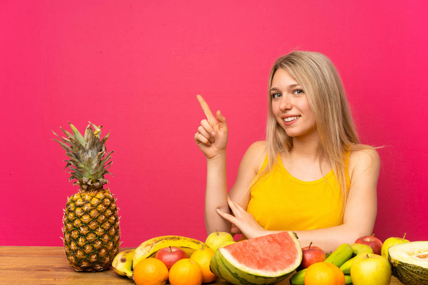 junge blonde Frau mit vielen Früchten zeigt mit dem Finger zur Seite - Foto, Bild