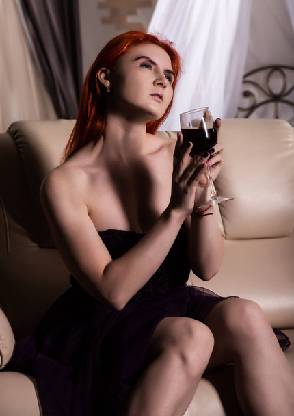 lonely young woman drinking wine in hotel room. redhead woman sitting on a sofa with glass of red wine - Fotoğraf, Görsel