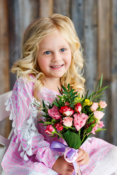 Beautiful little girl with blond hair and a bouquet of fresh flowers sits and smiles in a bright studio. Daylight - Zdjęcie, obraz