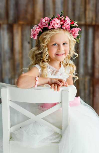 Lovely cute little girl - blonde in a wreath of live roses in a white beautiful dress in a bright studio. - Fotó, kép