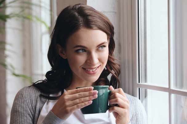 Όμορφη νεαρή γυναίκα πίνοντας τον ζεστό καφέ κοντά στο παράθυρο στο σπίτι - Φωτογραφία, εικόνα