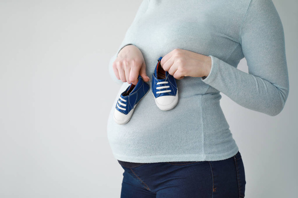 Hermosa mujer embarazada con botines de bebé sobre fondo claro
 - Foto, Imagen