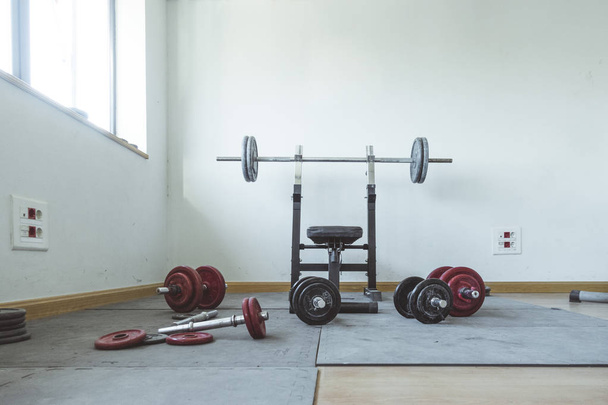 A sunlit weight area of a gym. There is a weight bench that is surrounded by stacks of weights. - Fotografie, Obrázek
