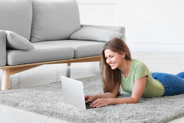 自宅で柔らかいカーペットの上に横たわっているラップトップを持つ若い女性 - 写真・画像