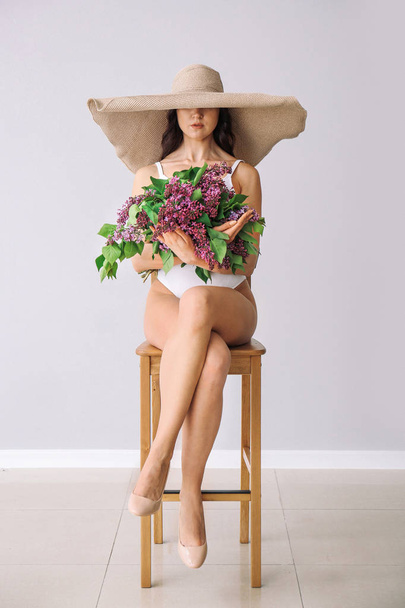 Beautiful young woman in swimming suit and with bouquet of lilac flowers sitting on chair against white wall - Фото, зображення