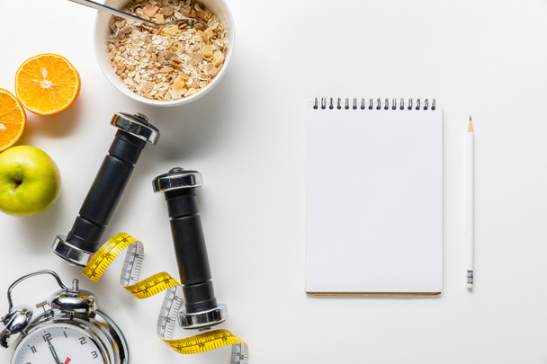 top view of dumbbells, measuring tape, alarm clock and breakfast cereal with fruits near empty notebook on white background - Valokuva, kuva