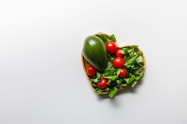 Κορυφαία θέα από φρέσκα πράσινα λαχανικά σε σχήμα καρδιάς μπολ σε λευκό φόντο - Φωτογραφία, εικόνα