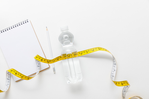 top view of water in bottle, measuring tape, blank notebook with pencil on white background  - Photo, Image