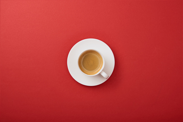Top uitzicht van witte beker met koffie op de schotel op rode achtergrond - Foto, afbeelding