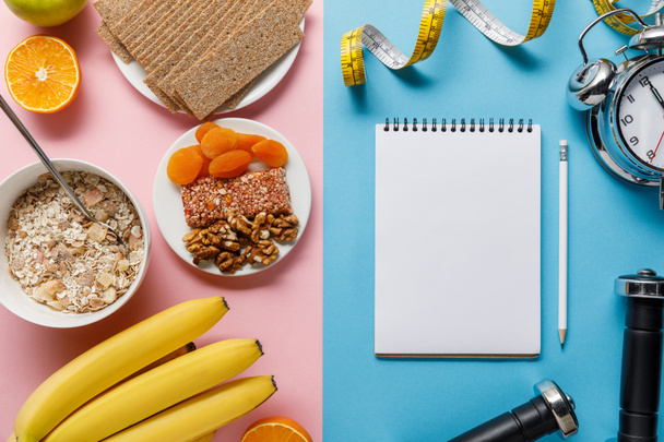 top view of fresh fruits, crispbread and breakfast cereal on pink and blank notebook, dumbbells and measuring tape on blue background  - Foto, Imagen