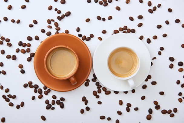 верхний вид кофе в белых и коричневых чашки на тарелках возле разбросанных жареных бобов
 - Фото, изображение