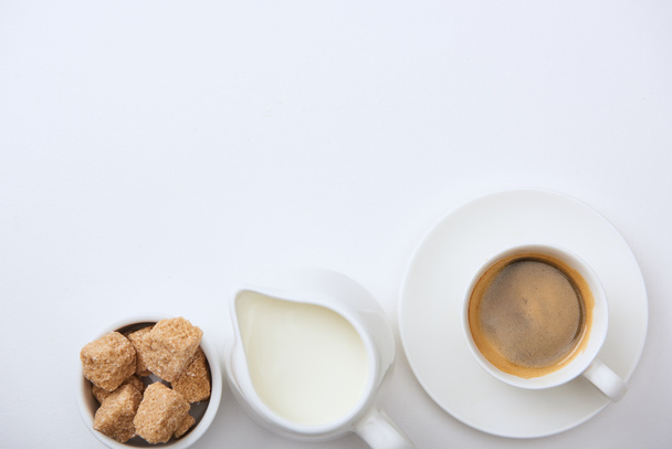 ylhäältä näkymä maukasta kahvia kuppi lautasella lähellä maitoa ja ruskeaa sokeria valkoisella pohjalla
 - Valokuva, kuva