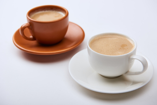 selective focus of delicious coffee with foam in cups on saucers on white background - Fotó, kép