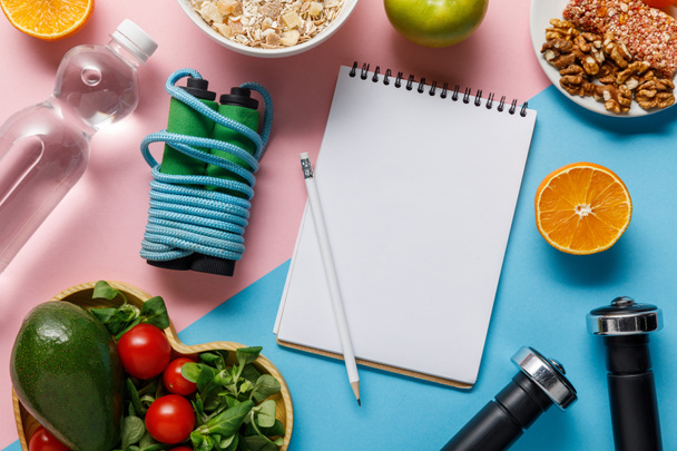 Draufsicht auf leeres Notizbuch und Bleistift mit leckerem Diätfutter, Wasser- und Sportgeräten auf blauem und rosa Hintergrund  - Foto, Bild