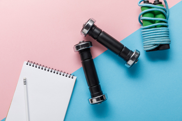 top view of skipping rope, dumbbells and notebook on blue and pink background - Fotoğraf, Görsel