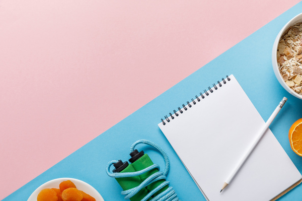 flat lay with empty notebook, skipping rope, breakfast cereal with orange and dried apricots on pink and blue background - Photo, Image