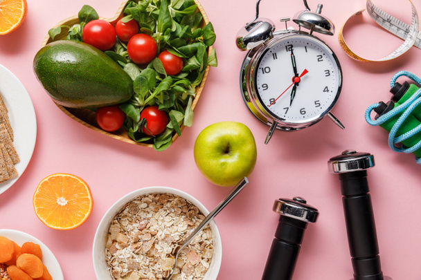 top view of fresh diet food, measuring tape, sport equipment and alarm clock on pink background with copy space - Photo, Image