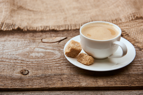 delicious coffee with foam in cup on saucer with brown sugar near sackcloth on wooden background  - Photo, Image