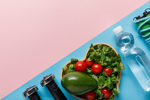 flat lay with vegetable salad in heart-shaped bowl, water, skipping rope and dumbbells on pink and blue background - Photo, Image