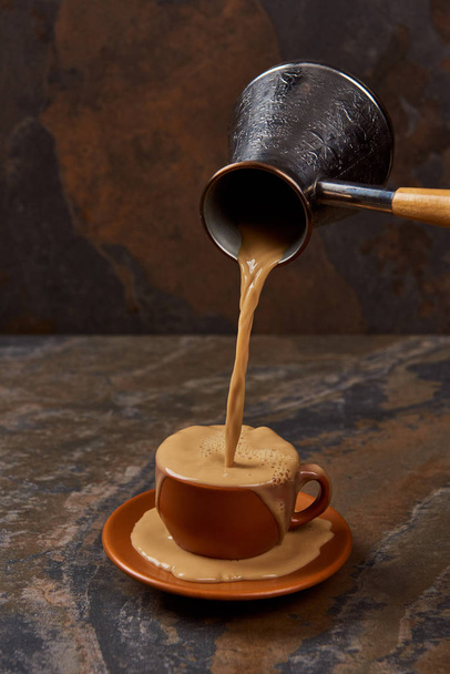 gieten van koffie van cezve in kop en op schotel op marmeren oppervlak - Foto, afbeelding
