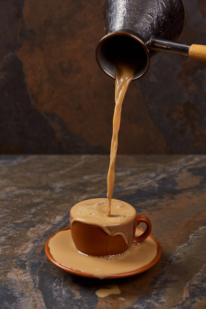pouring hot coffee from cezve into cup with puddle on saucer on marble surface - Photo, Image