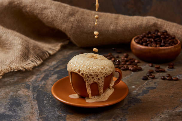 derramando café quente em xícara com espuma na superfície de mármore perto de pano de saco e feijão
 - Foto, Imagem