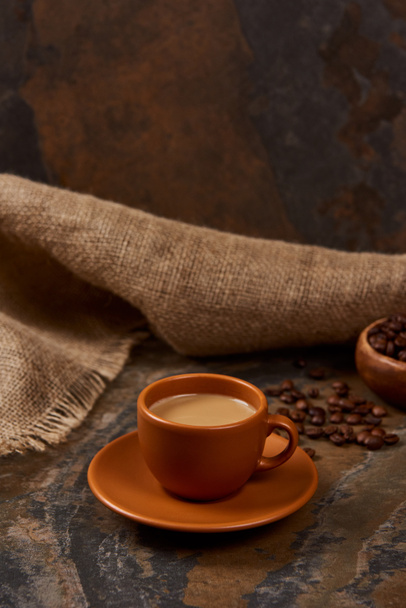 heißer Kaffee in Tasse auf Untertasse auf Marmoroberfläche in der Nähe von Sacktuch und Bohnen - Foto, Bild