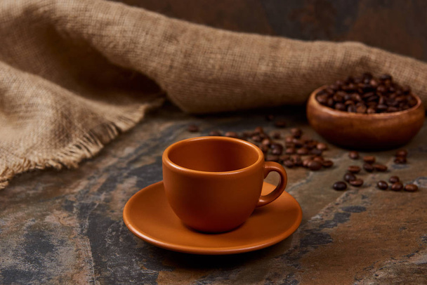 коричнева чашка на блюдці на мармуровій поверхні біля мішковини та кавових зерен
 - Фото, зображення