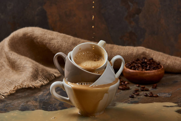 stroomt uit hete koffie uit kopjes met plas op marmeren oppervlak in de buurt van zak en bonen - Foto, afbeelding
