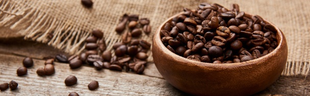 vista de perto de grãos de café torrados em tigela perto de pano de saco em tábua de madeira, tiro panorâmico
 - Foto, Imagem
