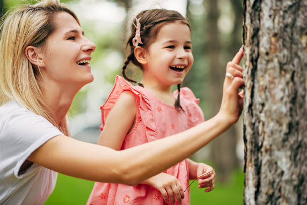 Outdoor portrait of happy young mother exploring with her cute daughter the nature in the park. Happy family relationship. Woman playing and laughig with her child outdoor exploring the life of nature - Photo, image