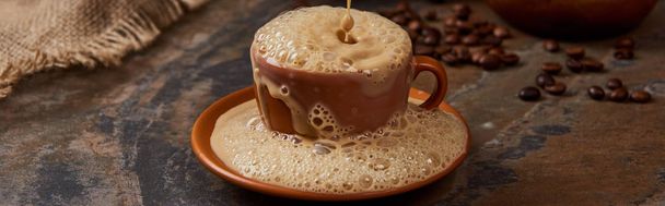 panoramic shot of coffee foam with bubbles pouring out of cup on saucer on marble surface  - Фото, изображение