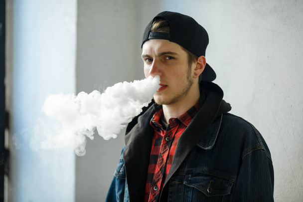 Vape teenager. Portrait of young handsome guy in a cap smoking an electronic cigarette on the street in the spring. Bad habit that is harmful to health. Vaping activity. Close up. - 写真・画像