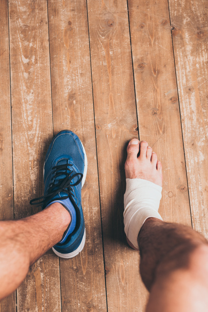 μερική θέα του τραυματισμένου αθλητή που κάθεται σε ένα παπούτσι με ένα πόδι σε ελαστικό επίδεσμο - Φωτογραφία, εικόνα