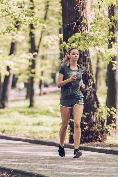 pretty young sportsman running and park, holding smartphone and listening music in earphones - Photo, Image