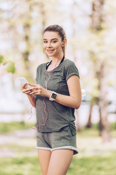 atrakcyjna kobieta w odzieży sportowej przy użyciu smartfona i słuchania muzyki w słuchawkach podczas uśmiechu w aparacie  - Zdjęcie, obraz