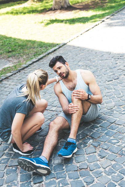 young woman in sportswear touching shoulder of injured sportsman sitting on pavement and suffering from pain - Foto, Imagen