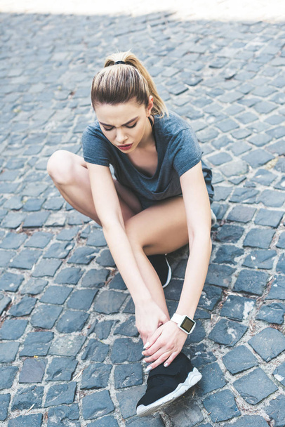 attractive sportswoman sitting on pavement and touching injured leg - 写真・画像