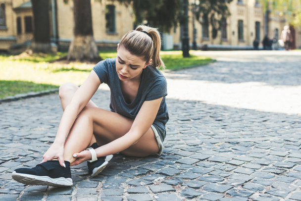 young pretty sportswoman sitting on pavement and touching injured leg  - Photo, Image