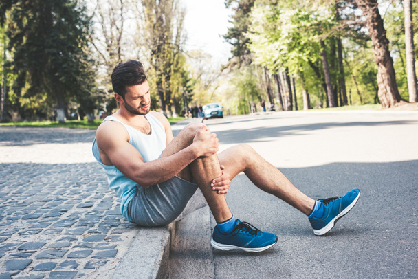 young sportsman sitting on pavement border and touching injured leg - Fotografie, Obrázek