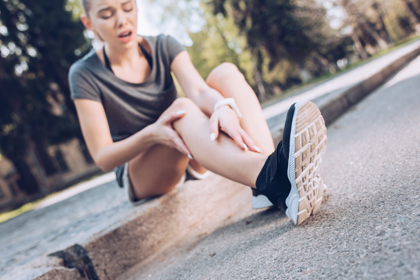 selective focus of young sportswoman sitting on border and touching injured leg - Фото, изображение