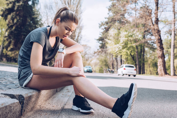 KYIV, UKRAINE - APRIL 25, 2019: Young sportswoman suffering from pain while sitting on pavement and touching injured leg.  - Photo, Image