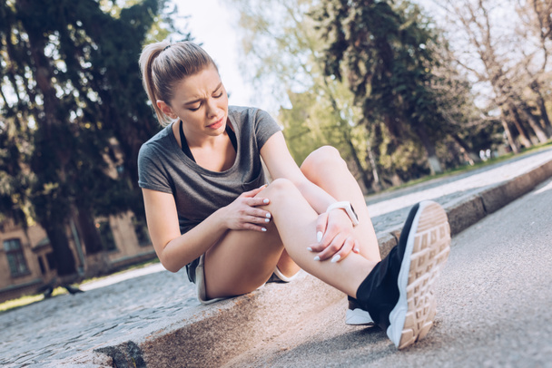 sad sportswoman suffering from pain while sitting on pavement and touching injured leg - Fotoğraf, Görsel