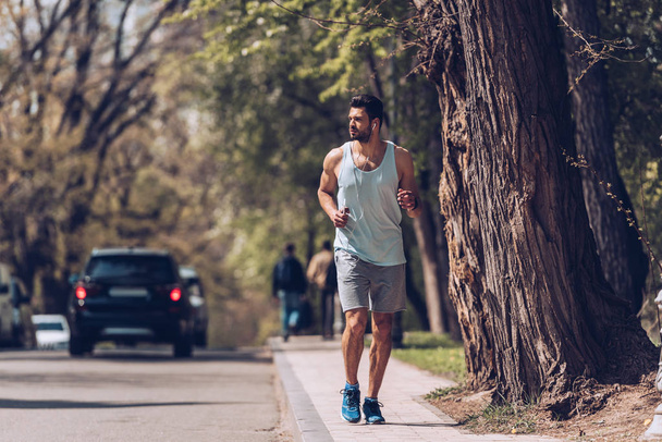 KYIV, UCRANIA - 25 DE ABRIL DE 2019: Guapo deportista corriendo por el pavimento cerca de la carretera y escuchando música en los auriculares
.  - Foto, imagen