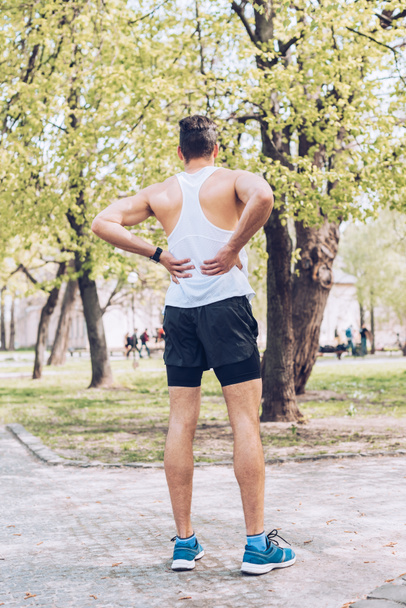 back view of man in sportswear and sneakers standing in park and touching injured back - Foto, Imagen