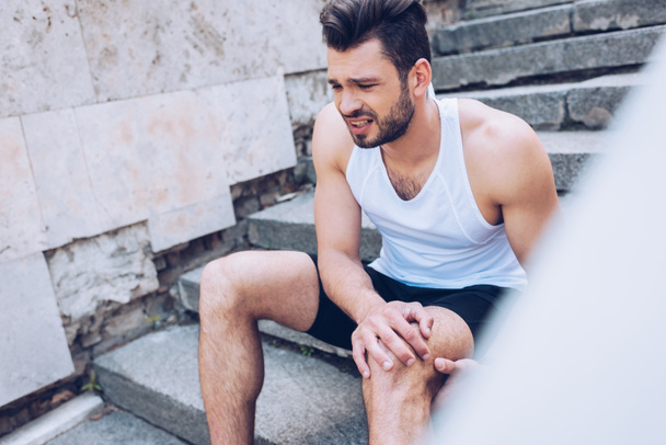 selective focus of handsome sportsman suffering from hurt while sitting on stairs and touching injured knee - Fotoğraf, Görsel