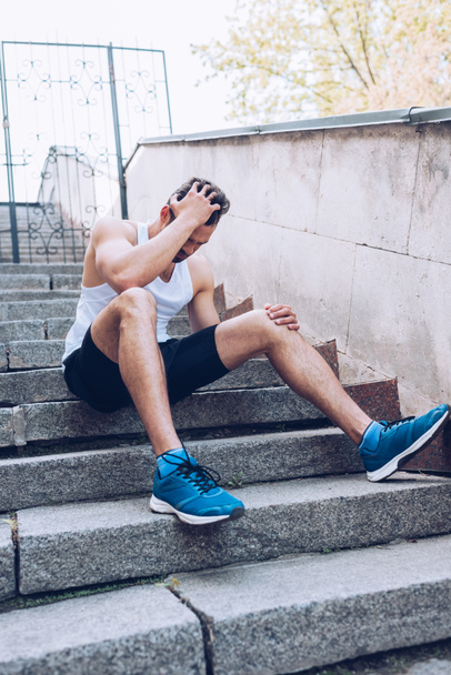 injured sportsman sitting on stairs and suffering from from pain while holding hand on head - Foto, imagen